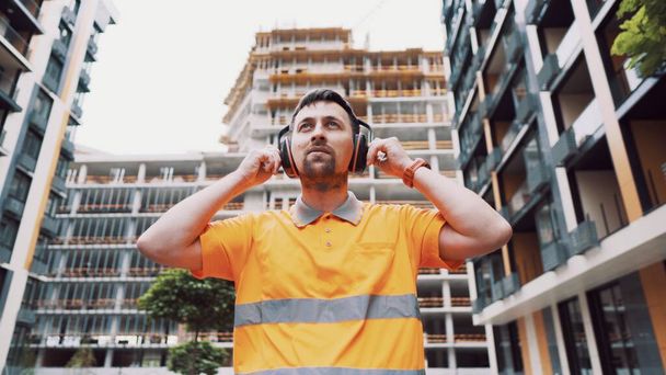Builder covers his ears, ear muff to protect workers ears. Construction worker wearing protective ear defenders. Concept of construction, taking care of safety during work. Protection against injury. - Photo, Image