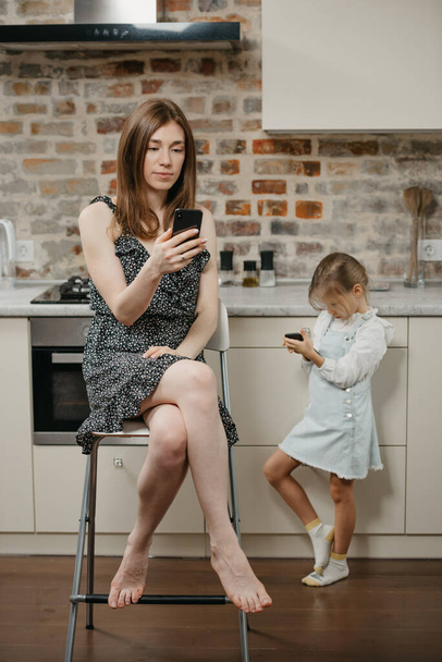 A mother with pale skin is reading news on a smartphone while sitting on a chair near a daughter which is gaming on a phone in the kitchen. A mom and her young girl are using their cellphones at home. - 写真・画像