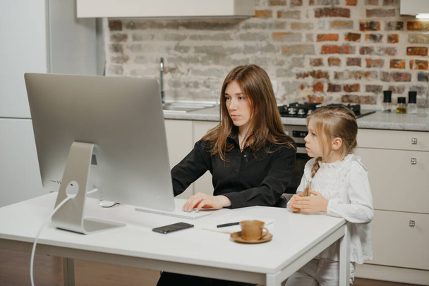 A mother is working remotely at home while a daughter is watching it. A businesswoman is working from an apartment near her blonde child. - Foto, Imagen