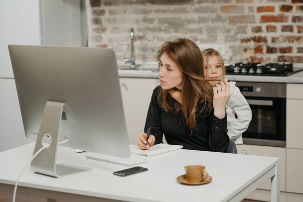 A young mother is working remotely at home while a daughter is hugging her. A businesswoman is doing notes while working from an apartment near her blonde child. - 写真・画像