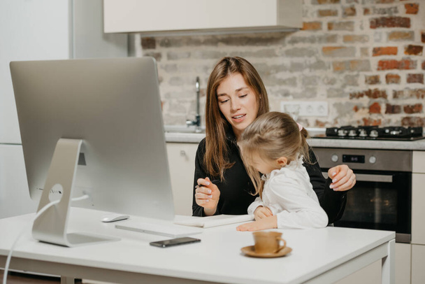 A young mother is describing to a daughter what is she doing. A smiling businesswoman is taking notes while working from an apartment near her blonde child. - Photo, Image