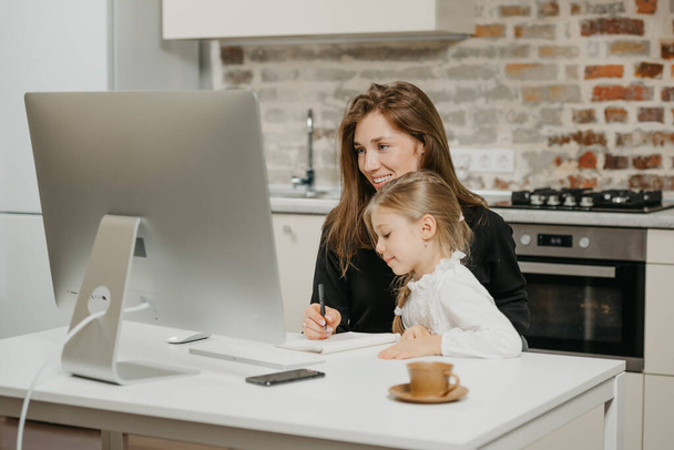 Mladá matka pracuje na dálku doma, zatímco dcera studuje u ní. Usmívající se podnikatelka si dělá poznámky, když pracuje z bytu poblíž svého blonďatého dítěte. - Fotografie, Obrázek