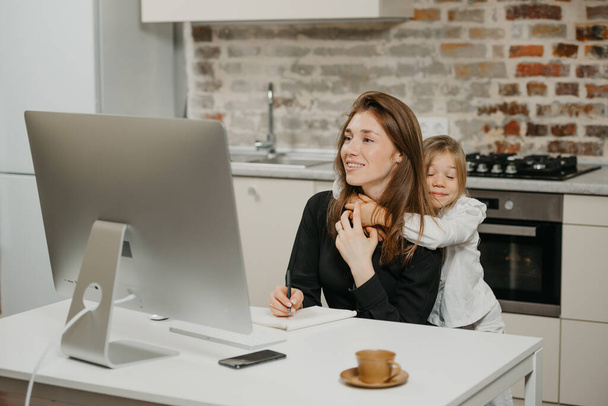 A young mother is working remotely at home while a daughter is hugging her. A smiling businesswoman is taking notes while working from an apartment near her blonde child. - Photo, Image