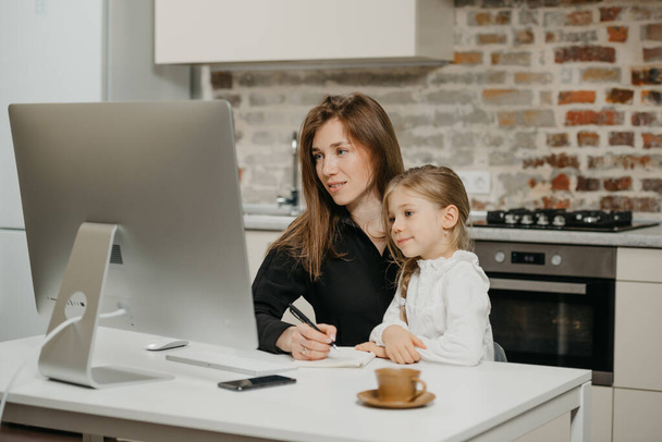 Uma jovem mãe está trabalhando remotamente em casa, enquanto uma filha feliz está olhando para um computador. Uma mãe bonita está tomando notas enquanto trabalhava em um apartamento perto de seu filho loiro. - Foto, Imagem