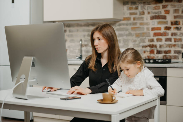 Młoda mama pracuje zdalnie, podczas gdy córka rysuje w notesie w domu. Piękna matka czyta wiadomości na komputerze w pobliżu swojego blond dziecka, które pilnie pisze. - Zdjęcie, obraz