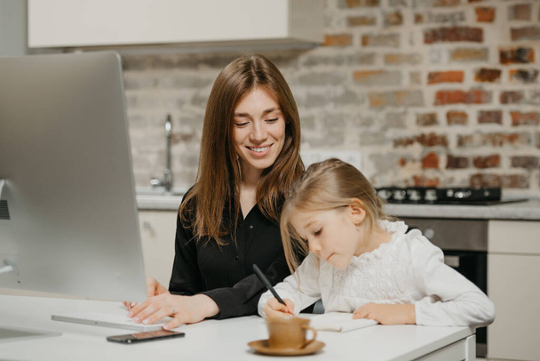 Uśmiechnięta mama gapi się na córkę, która rysuje w notatniku w domu. Ładna matka pracuje zdalnie przy komputerze w pobliżu swojego blond dziecka, które pilnie pisze listy.. - Zdjęcie, obraz