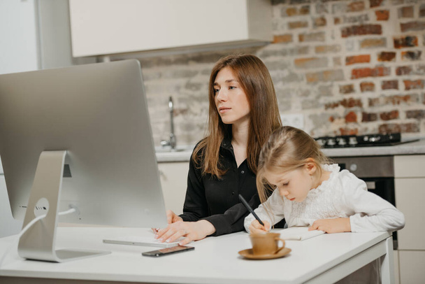 Uma jovem mãe está trabalhando remotamente com cuidado enquanto uma filha está desenhando o caderno em casa. Uma mãe linda está estudando em um computador perto de seu filho loiro que está escrevendo cartas diligentemente. - Foto, Imagem