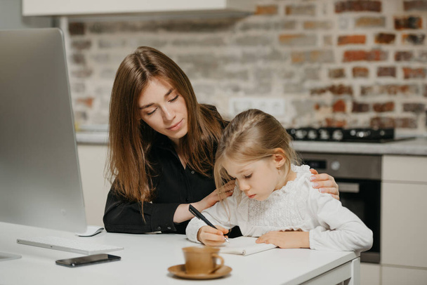 A young mom is hugging a pretty daughter while she is drawing in the notebook at the workplace. A gorgeous mother is looking at her blonde child which is doing homework at home. - Photo, Image