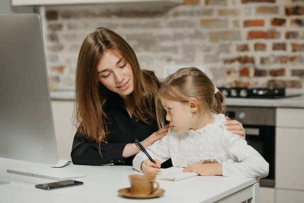 A young smiling mom is hugging a pretty daughter while she is drawing in the notebook at home. A gorgeous mother is looking at her blonde child which is doing homework. - Photo, Image