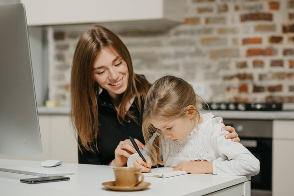 Een jonge lachende moeder knuffelt een mooie dochter terwijl ze op het werk in het notitieboekje schrijft. Een prachtige moeder is op zoek naar haar blonde kind die huiswerk thuis doet. - Foto, afbeelding