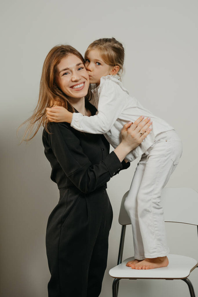 Мама обіймає доньку, яка стоїть на барному стільці. Усміхнена мама і дитина позують
 - Фото, зображення
