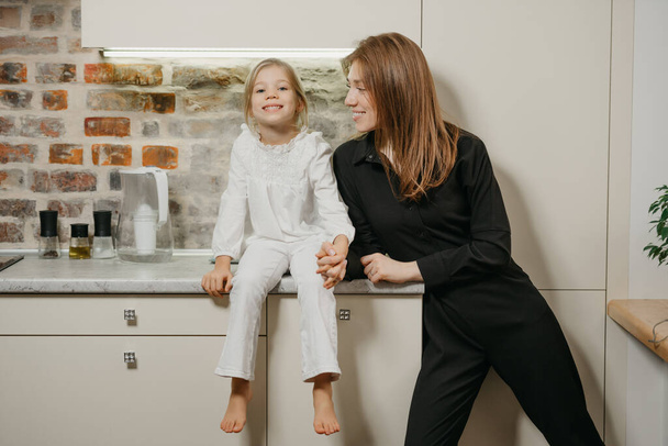 Een blanke jonge moeder in een zwart jumpsuit kijkt naar haar mooie dochter in de keuken. Een lachende moeder staat, haar gelukkige meisje zit thuis op een aanrecht. - Foto, afbeelding