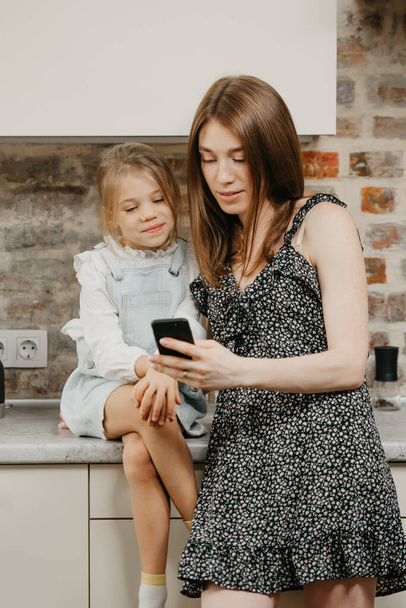 A caucasian young mother with pale skin is reading the news on a phone with a daughter in the kitchen. A mom is using a cellphone while her happy pretty girl is sitting on a table. - Valokuva, kuva