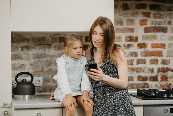 Eine junge europäische Mutter mit blasser Haut liest die Nachrichten am Telefon mit einer Tochter in der Küche. Eine Mutter benutzt ein Handy, während ihr hübsches Mädchen auf einer Arbeitsplatte sitzt. - Foto, Bild