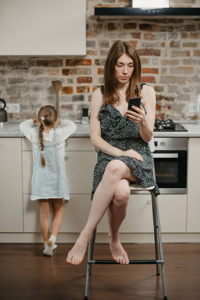 Een blanke moeder met een bleke huid leest nieuws op een smartphone terwijl ze op een stoel zit in de buurt van een dochter die zich van haar afkeert in de keuken. Een moeder gebruikt een mobieltje in de buurt van haar verveelde kind thuis. - Foto, afbeelding