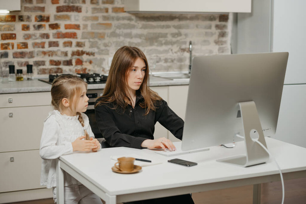 A mother is working remotely at home while a daughter is watching it. A businesswoman is working from an apartment near her blonde child. - Photo, Image