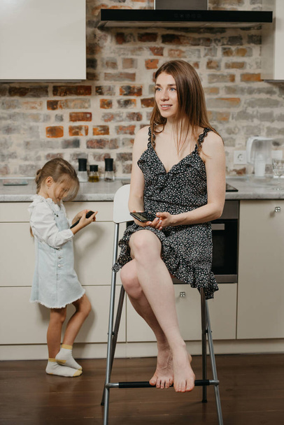 Hymyilevä äiti mekossa haaveilee istuessaan tuolilla lähellä tytärtä, joka pelaa puhelimessa keittiössä. Äiti ja nuori tyttö käyttävät kännyköitä kotona.. - Valokuva, kuva