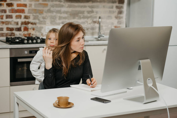 Egy fiatal anya távolról dolgozik otthon, miközben a lánya ölelgeti. Egy üzletasszony jegyzetel, miközben egy lakásban dolgozik a szőke gyermeke mellett.. - Fotó, kép