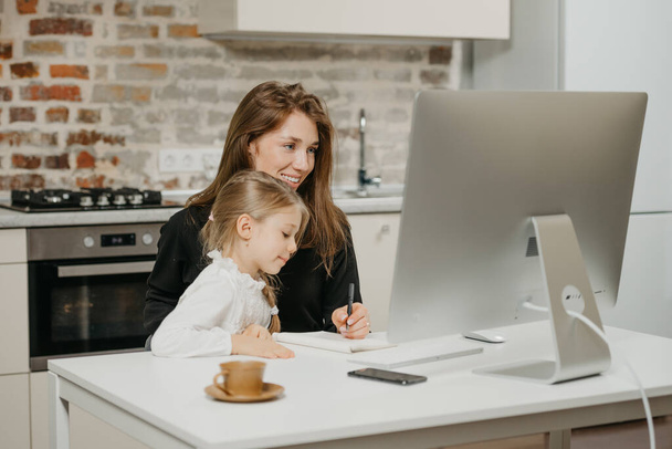 A young mother is working remotely at home while a daughter is studying from her. A smiling businesswoman is taking notes while working from an apartment near her blonde child. - Photo, Image