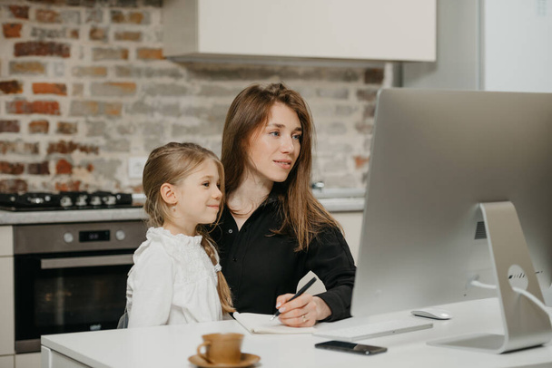 Молодая мама удаленно работает дома, в то время как счастливая дочь смотрит на компьютер. Красивая деловая женщина делает заметки во время работы в квартире рядом со своим блондинкой ребенком. - Фото, изображение