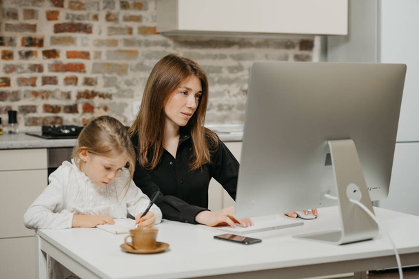 Młoda mama pracuje zdalnie, podczas gdy córka rysuje w notesie w domu. Piękna matka uczy się na komputerze w pobliżu swojego blond dziecka, które pilnie pisze. - Zdjęcie, obraz