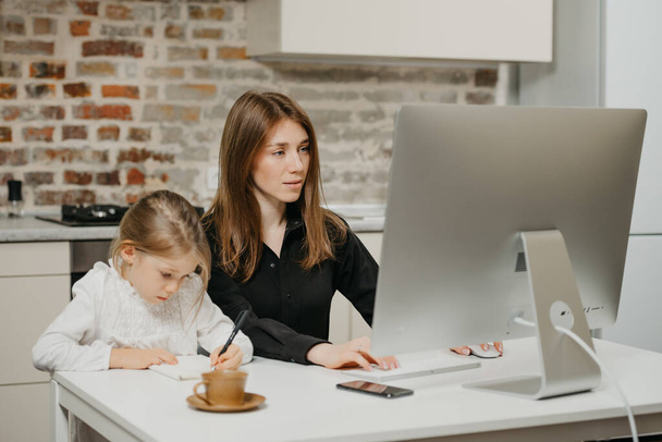 Μια νεαρή μαμά εργάζεται εξ αποστάσεως προσεκτικά, ενώ μια κόρη ζωγραφίζει στο σημειωματάριο στο σπίτι. Μια όμορφη μητέρα μελετάει σε έναν υπολογιστή κοντά στο ξανθό παιδί της που γράφει γράμματα επιμελώς.. - Φωτογραφία, εικόνα