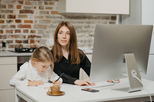 A young mom is staring away while a daughter is drawing in the notebook at home. A pretty mother is working remotely on a computer near her child which is writing letters diligently. - Photo, Image