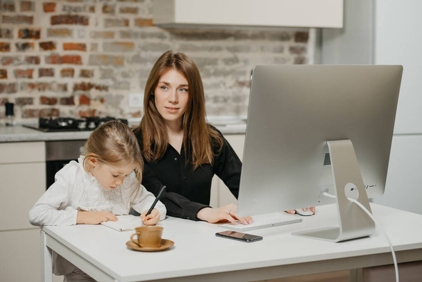 A mom is staring away while a daughter is drawing in the notebook at home. A pretty mother is working remotely on a computer near her child which is writing letters diligently. - Photo, Image