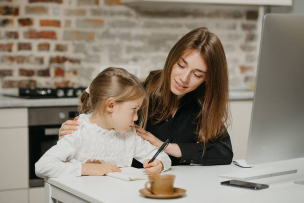 A young smiling mom is hugging a pretty daughter while she is drawing in the notebook at home. A gorgeous mother is looking at her blonde child which is doing homework. - Photo, Image