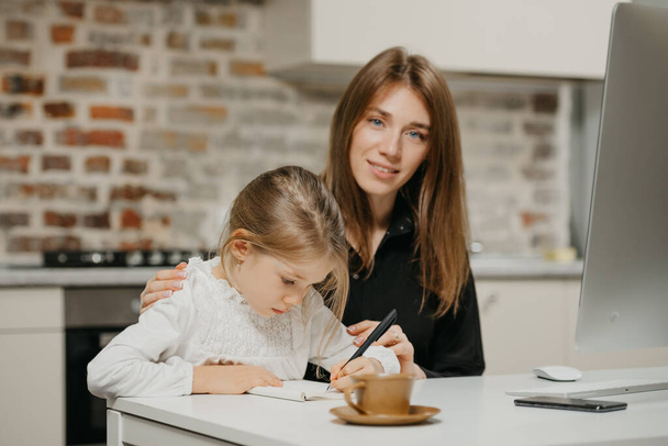 A mom is posing and hugging a pretty daughter while she is drawing in the notebook at the workplace. A gorgeous mother watches her blonde child which is doing homework at home. - Photo, Image