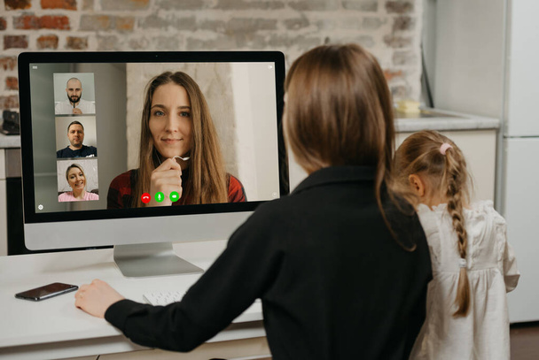 Une jeune mère lors d'une vidéoconférence avec des collègues sur l'ordinateur de bureau pendant que sa fille dessine. Une maman magnifique travaille à distance avec son enfant blond à la maison - Photo, image