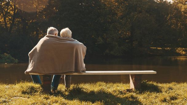 Ouder grijs harig echtpaar zittend op de bank in de buurt van de rivier bedekt met deken op herfstdag. Romantiek en ouderdom - Foto, afbeelding