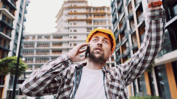 Caucasian male construction worker in orange hard hat and plaid shirt talking on phone at construction site. Architecture theme. Male profession. Foreman controls construction process by smartphone. - Фото, зображення