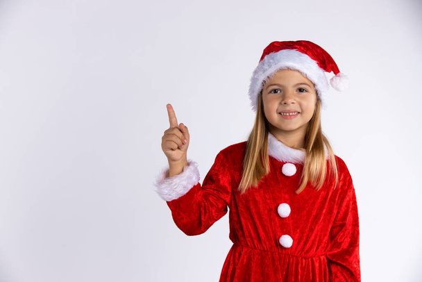  Belle petite fille habillée en Père Noël avec un chapeau d'hiver. Portrait d'une fille pointant son doigt vers le haut, isolée sur fond blanc avec espace libre - Photo, image