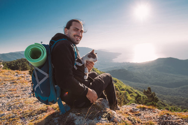 A male tourist meets a dawn in the mountains. Hiking. A young handsome guy climbed to the top of the mountain in the morning and sits on the edge of a cliff with his friend cat. - Photo, Image