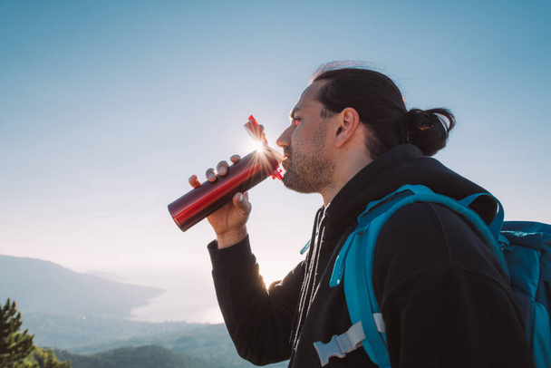 Un excursionista bebe té de un termo en la cima de una montaña al amanecer. Joven con equipo turístico se encuentra alegremente con el sol de la mañana en las montañas con vistas al mar - Foto, imagen