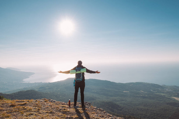 Turysta stoi z wyciągniętymi ramionami na szczycie góry o wschodzie słońca. Młody człowiek ze sprzętem turystycznym z radością wita poranne słońce w górach z widokiem na morze - Zdjęcie, obraz