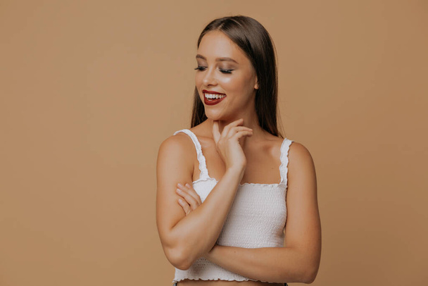 Elegant girl with beautiful brown eyes touching chin with fingers and gently smilling. Close-up portrait of trendy young woman in white t-shirt posing in studio. High quality photo - Foto, Imagen