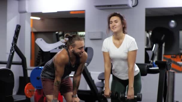 Gym Lektion mit lustigem Trainer Mann Anfängerin junge Frau schüttelt ihren Rücken nach vorne beugt sich mit Hanteln - Filmmaterial, Video