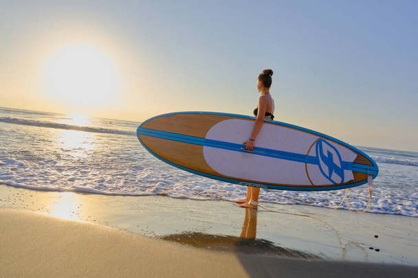 jovem menina bonita em um maiô com uma prancha de surf ao amanhecer - Foto, Imagem