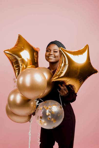 Muslim black girl with golden helium balloons smiles isolated pink background. African young woman celebrates her birthday - Фото, зображення
