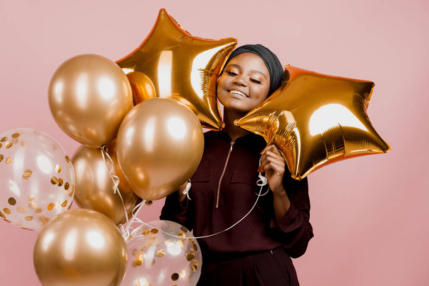 Christmas celebration. African american holiday. Attractive girl with golden helium balloons isolated peach background. Young black woman weared traditional dress and scarf. - Φωτογραφία, εικόνα
