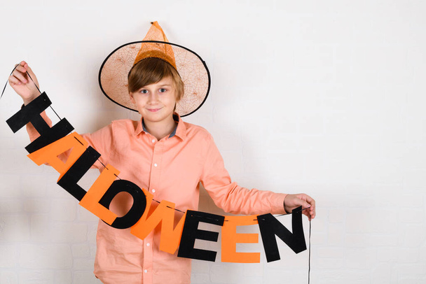 オレンジの帽子のブロンドの少年は、ハロウィンの碑文を掛け、白いレンガの壁の背景に自宅でハロウィンを祝うために彼の部屋を飾ります - 写真・画像