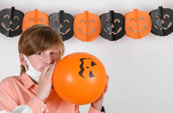 Um menino loiro em máscara médica protetora infla um balão laranja, decora seu quarto para celebrar o Halloween em um fundo de parede de tijolo branco. Feliz Halloween em quarentena coronavírus pandemia - Foto, Imagem
