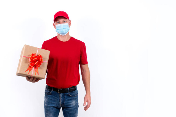 Safe delivery of gifts for holidays. A courier in red uniform and protective medical mask holds box with a bow. Contactless remote gift orders during pandemic. Banner with space for text. Copy space. - Photo, image