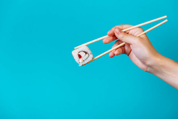 Woman's hand holds Tasty sushi roll maki with wooden chopsticks on blue background close up. Place for caption and text. - 写真・画像