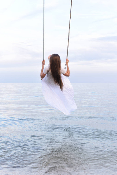 Piękna szczęśliwa dziewczyna w białej sukience jeździ na huśtawce liny nad morzem. Rama pionowa. Widok z tyłu. - Zdjęcie, obraz