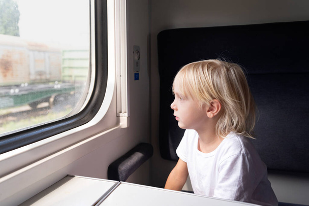 A szőke kisfiú figyelmesen néz ki a vonat ablakán. Vasúti utazás gyermekekkel. - Fotó, kép