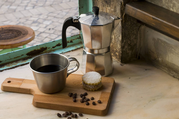 Ιταλικός μαύρος καφές σε ένα φλιτζάνι με κόκκους καφέ και ένα χειροποίητο μπισκότο με ζάχαρη και μια κατσαρόλα Moka πίσω. Τα πάντα στο παλιό καφέ φόντο κοντά στο παράθυρο. Υγιές πρωινό. - Φωτογραφία, εικόνα