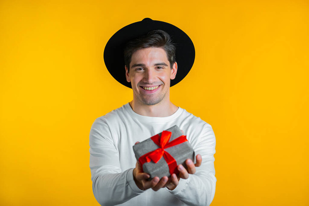 Excited man holding gift box and gives it by hands to camera on yellow wall background. Guy smiling, he is happy with present. Studio portrait - Foto, Imagen